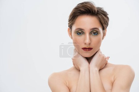 portrait of charming young woman with bright makeup and short hair touching neck isolated on grey