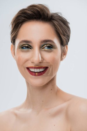 Porträt einer fröhlichen jungen Frau mit glänzendem Make-up und kurzen Haaren isoliert auf grau 
