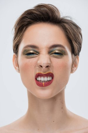 Porträt einer emotionalen jungen Frau mit glänzendem Make-up und kurzen Haaren, die vereinzelt auf grau grinsen 