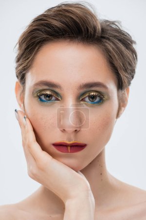 portrait of young woman with shiny makeup and short hair holding hand near face isolated on grey 