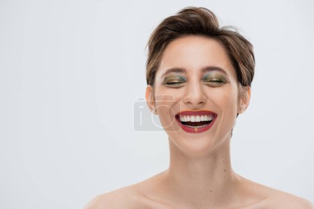 Foto de Mujer joven positiva con maquillaje brillante y pelo corto sonriendo aislado en gris - Imagen libre de derechos