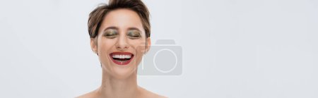 positive young woman with shiny makeup and short hair smiling isolated on grey, banner 