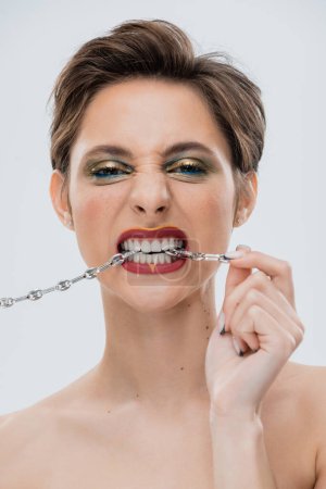 portrait of young woman with bright makeup and short hair biting silver chain isolated on grey 