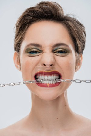 Foto de Retrato de mujer joven con pelo corto y labios rojos mordiendo cadena de plata aislada en gris - Imagen libre de derechos