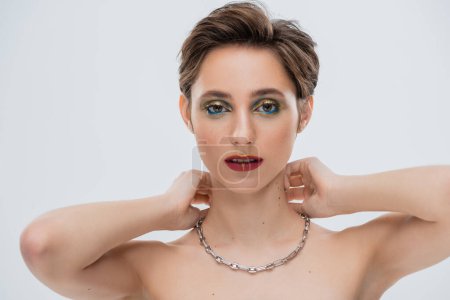 young woman with shiny makeup and short hair wearing silver chain necklace isolated on grey 