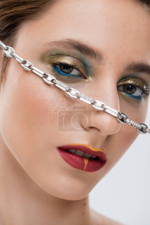Nahaufnahme einer jungen Frau mit glänzendem Augen-Make-up und silberner Kette im Gesicht isoliert auf grau 