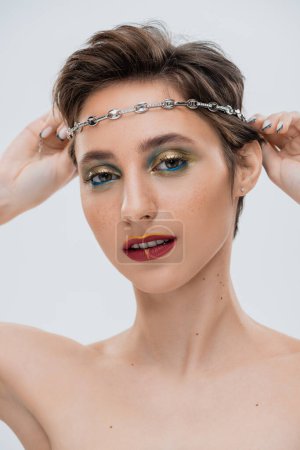 young woman with shiny makeup and short hair wearing silver chain on head isolated on grey 