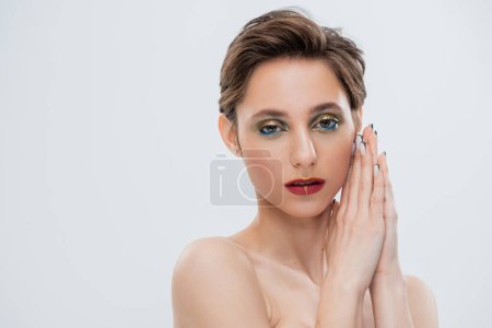 young woman with shimmery eye makeup and short hair standing with praying hands isolated on grey 