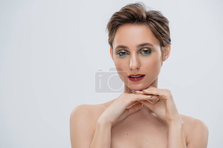 junge Frau mit schimmerndem Augen-Make-up und kurzen Haaren blickt vereinzelt in die Kamera auf grau 