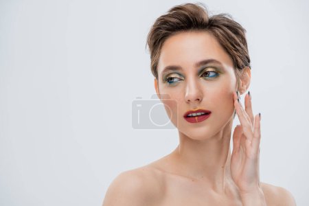 junge Frau mit schimmerndem Augen-Make-up und kurzen Haaren, die die Wange berühren, isoliert auf grau 