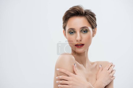 young woman with shimmery eye makeup and short hair standing with crossed arms isolated on grey 