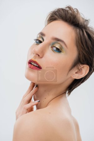 brunette jeune femme avec maquillage des yeux chatoyant et cheveux courts touchant cou isolé sur gris 