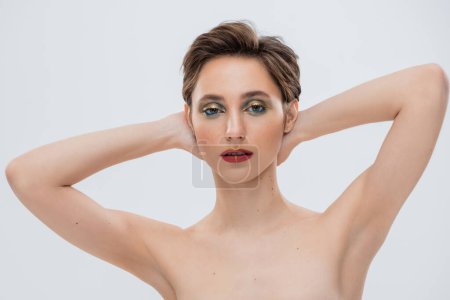 young woman with shimmery eye makeup standing with hands behind neck isolated on grey 