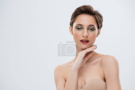 pretty young woman with shimmery eye makeup touching chin isolated on grey 