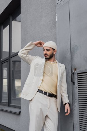 Fashionable homosexual man in beige suit and hat looking away near building outdoors 