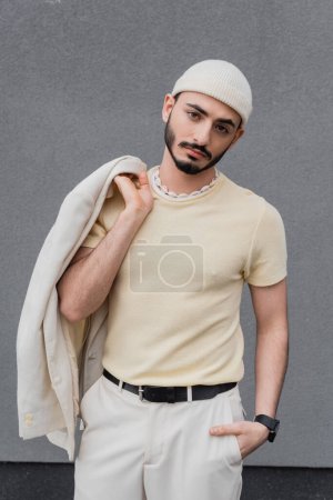 Téléchargez les photos : Portrait d'homme gay à la mode en chapeau tenant veste et posant à l'extérieur - en image libre de droit