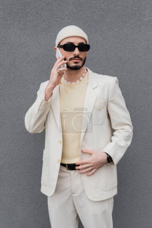 Hombre gay de moda en traje y gafas de sol hablando en el teléfono inteligente cerca del edificio al aire libre 
