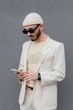 Hombre gay de moda en gafas de sol usando teléfono inteligente cerca de la pared al aire libre 