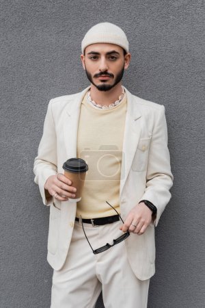 Fashionable gay man in suit holding coffee to go and sunglasses near wall outdoors 