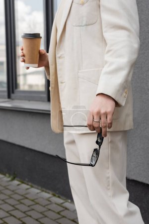 Cropped view of gay man in suit holding sunglasses and coffee to go outdoors 
