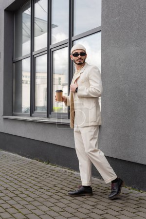 gay hombre en elegante traje y gafas de sol celebración café a ir en urbano calle 