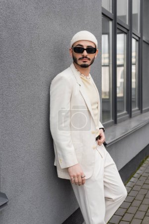 Stylish homosexual man in sunglasses and suit standing near facade of building outdoors 