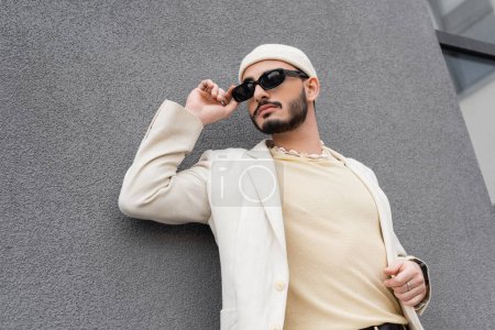 Hombre gay bien vestido en chaqueta y sombrero tocando gafas de sol en la calle urbana 