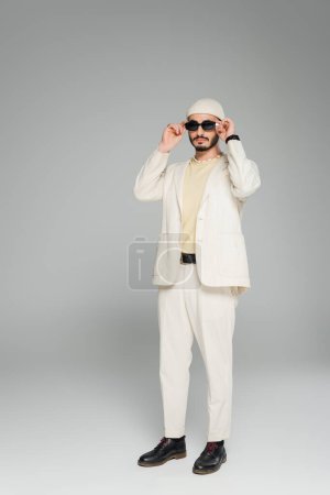 Volle Länge des trendigen homosexuellen Mannes in beigem Anzug und Hut mit Sonnenbrille auf grauem Hintergrund 