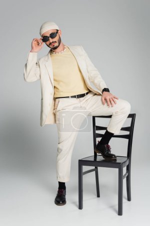 Hombre gay de moda en traje y gafas de sol posando con silla sobre fondo gris 