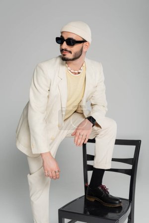 Fashionable and bearded gay man in suit standing near chair isolated on grey