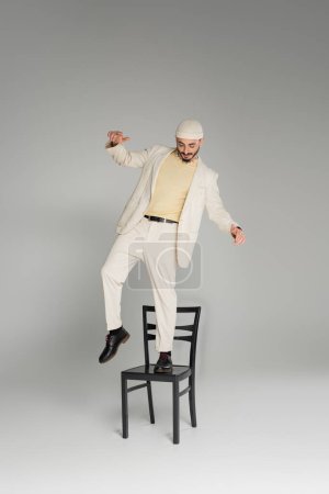 Stylish gay man in beige suit and hat standing on chair on grey background 