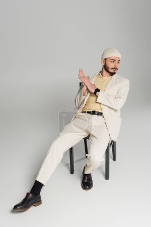 Trendy gay man in beige hat and suit sitting on chair on grey background 