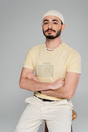 Porträt eines trendigen Schwulen mit Hut, der die Arme in der Nähe eines Stuhls kreuzt, isoliert auf grau  