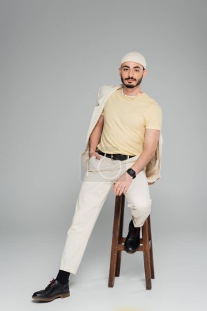 Well dressed gay man in suit and hat posing on chair on grey background 