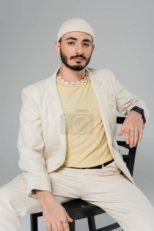 Photo for Bearded homosexual man in suit ad seashell necklace sitting on chair isolated on grey - Royalty Free Image
