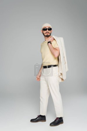 Stylish gay man in sunglasses holding jacket while standing on grey background 