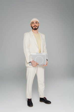 Stilvoller schwuler Mann im beigen Anzug mit Laptop auf grauem Hintergrund 