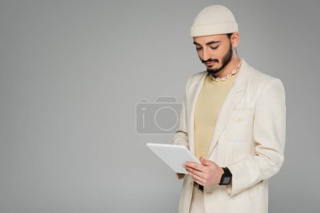 Young gay man in beige suit and hat using digital tablet isolated on grey  