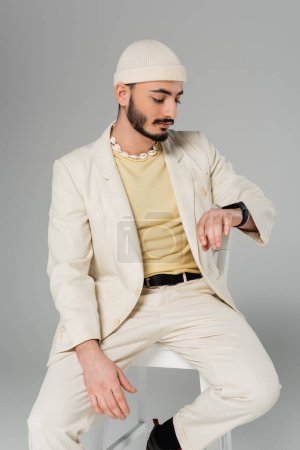 Foto de Elegante gay hombre en traje mirando inteligente reloj mientras sentado en silla aislado en gris - Imagen libre de derechos