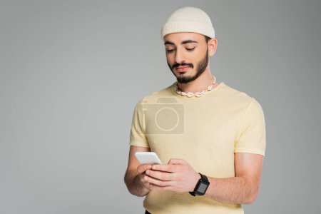 El hombre gay de moda en sombrero usando el teléfono celular aislado en gris  