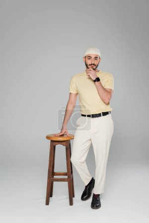 Stilvoller homosexueller Mann mit Hut und smarter Uhr steht neben Stuhl auf grauem Hintergrund 