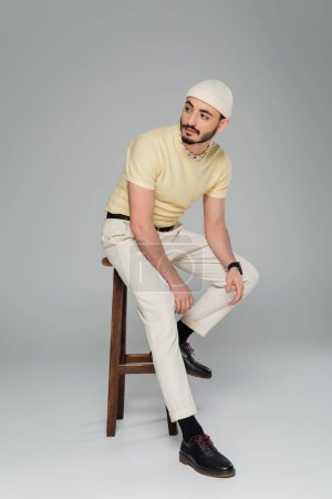 Foto de Longitud completa de moda gay hombre en beige sombrero sentado en silla en gris fondo - Imagen libre de derechos