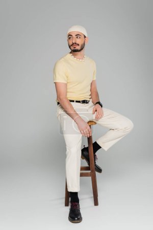 Téléchargez les photos : Jeune homme homosexuel en chapeau beige regardant loin tout en étant assis sur une chaise sur fond gris - en image libre de droit