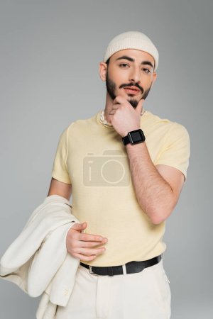 Téléchargez les photos : Portrait d'homme gay élégant en chapeau tenant veste beige et menton touchant isolé sur gris - en image libre de droit