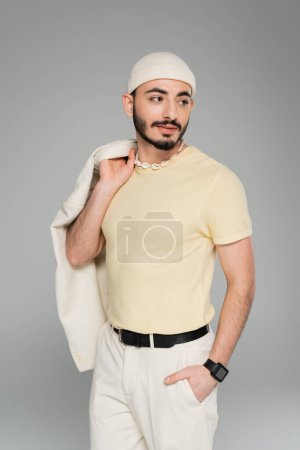 Portrait of stylish gay man in beige hat posing and holding jacket isolated on grey  