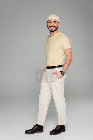 Full length of cheerful gay man in hat posing on grey background 
