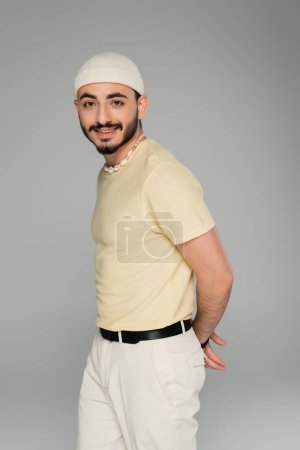 Porträt eines trendigen homosexuellen Mannes mit Hut und Muschelkette, der isoliert auf grau in die Kamera lächelt  