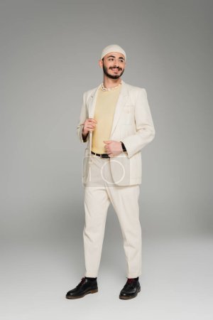 Cheerful homosexual man in beige suit and hat touching jacket on grey background 