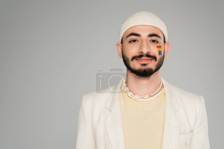 Retrato de un hombre gay con estilo con bandera lgbt en la mejilla mirando a la cámara aislada en gris  