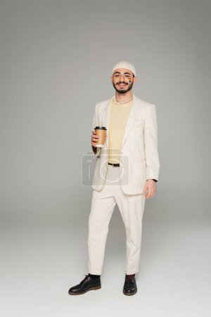 Cheerful gay man in suit with lgbt flag on cheek holding paper cup on grey background 